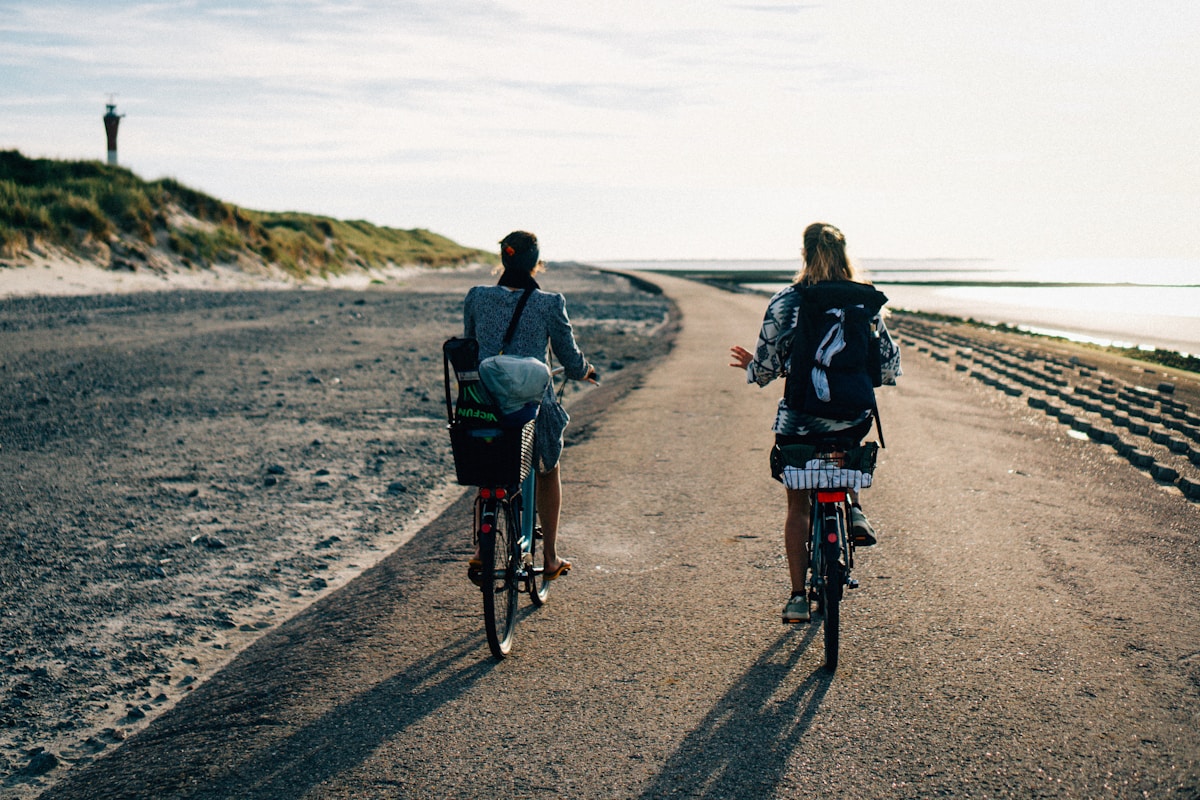Two cyclists seen from behind riding on a sunny paved path between grassy dunes and the ocean with a distant lighthouse